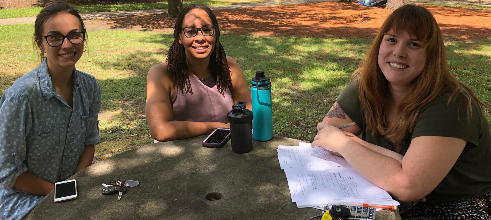 三个微笑, female, 白人学生围坐在一起, grey stone table: the first has brown hair with blond highlights worn down and is wearing a black shirt and a black skirt with multi-colored polka dots and is pictured with a stack of paper under her crossed arms on the table with a ring of keys including yellow and white cards and a black cell phone; the second has brown hair worn down and black rimmed glasses with a lavendar pink shirt, 她和一个高个子的合照, 蓝绿色和黑色的水瓶, 一个较短的黑色固体水瓶, and her cell phone with a lavendar back and white edges; 和 third has on a blue, 白色圆点领衬衫和棕色裤子, 照片中她拿着车钥匙和白色的手机, 面部上下部分的横杠.